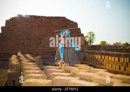 Eine unabhängige, schöne, hart arbeitende Frau, die auf dem Ziegelsteinofen-Gelände arbeitet. Aktiver traditioneller Ziegelofen im Hintergrund. Stockfoto