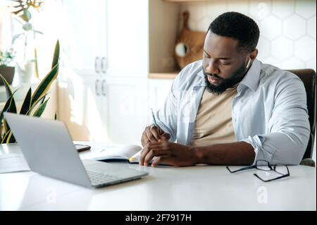 Konzentrierter, erfolgreicher, ernsthafter afroamerikanischer Bärtiger, Geschäftsleiter, Manager-Student, in stilvoller Kleidung, arbeitet oder studiert fernab zu Hause, benutzt einen Laptop, sitzt an einem Tisch, notiert Stockfoto