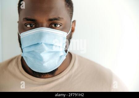 Nahaufnahme eines afroamerikanischen Mannes mit einer schützenden medizinischen Maske auf seinem Gesicht, der direkt auf die Kamera schaute, sich im Haus aufhielt und wegen Quarantänebeschränkungen isoliert blieb Stockfoto