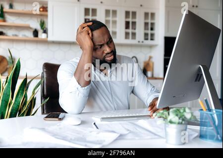 Verärgert trauriger junger afroamerikanischer Mann, Manager, Makler oder Freiberufler, der zu Hause am Computer arbeitet, Stress bei der Arbeit erlebt, seinen Kopf mit der Hand hält und traurig auf den Bildschirm schaut Stockfoto