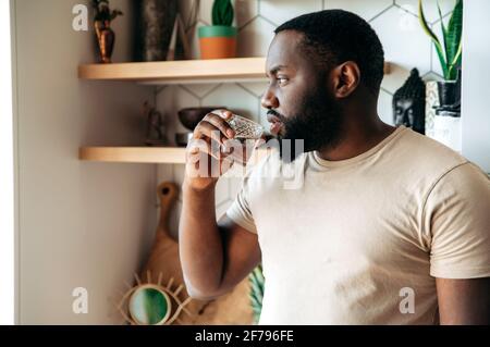Gesunder Lebensstil. Gutaussehender nachdenklicher gesunder junger afroamerikanischer bärtiger Mann, steht in lässiger Kleidung in der Küche, trinkt sauberes Wasser aus einem Glas, schaut zur Seite Stockfoto