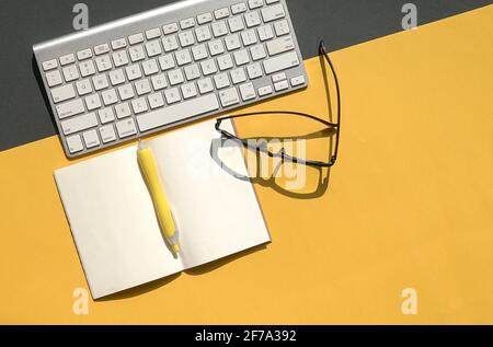 Kabellose Tastatur, Lesebrille und offener Notizblock auf schwarzem und gelbem Hintergrund oben. Speicherplatz kopieren. Stockfoto