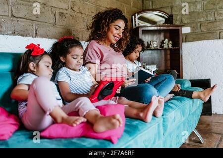 Junge lateinerin und ihre kleinen Kinder lesen bei Zuhause in Mexiko-Stadt Stockfoto
