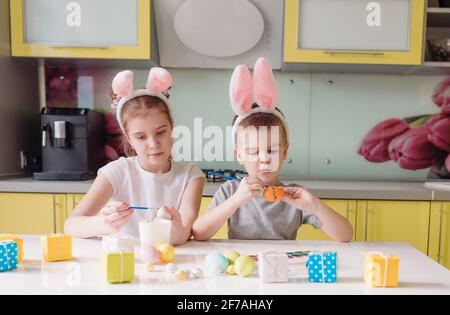 Ein kleiner Junge und ein Mädchen mit Kaninchenohren auf dem Kopf malen Ostereier, die zu Hause in der Küche sitzen Stockfoto