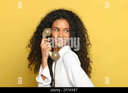 Glücklich lächelnde schwarze Frau hält einen retro Telefonhörer als Sie blickt über ihre Schulter auf ein gelbes Studio Hintergrund in einer Nahaufnahme Kopf und Stockfoto