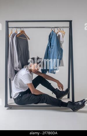 Lockiger junger Mann, der neben den Kleiderbügeln mit Kleidung und sitzt Sieht müde aus Stockfoto