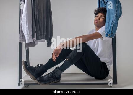 Lockiger junger Mann, der neben den Kleiderbügeln mit Kleidung und sitzt Sieht müde aus Stockfoto