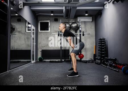 Totes Lifting-Training mit einer Kesselglocke. Der Mann in Sportbekleidung mit einem schwarzen T-Shirt steht mit gespreizten Beinen und hebt mit beiden Armen eine Kettelglocke Stockfoto