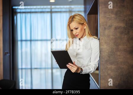 Konzentriert auf die Arbeit. Eine hübsche junge Geschäftsfrau, formell und hübsch gekleidet, nutzt ein digitales Tablet, während sie in Innenräumen auf einem modernen Hintergrund steht. Stockfoto