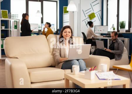 Glückliche Managerin, die auf dem Sofa vor der Kamera sitzt und lächelnd den Laptop hält, während verschiedene Kollegen im Hintergrund arbeiten. Multiethnische Mitarbeiter analysieren Finanzberichte von Start-ups in modernen Büros Stockfoto