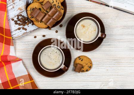 Zwei Tassen Kaffee mit Schokoladenkeksen und Schokolade auf dem Hintergrund eines offenen Buches. Heißer Kaffee mit Schokoladenkeksen im Tee Stockfoto