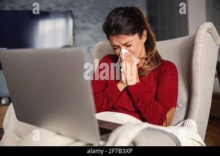 Kranke Frau, die zu Hause auf einem Stuhl sitzt, während sie einen Laptop im Schoß hatte und die Nase blies. Stockfoto