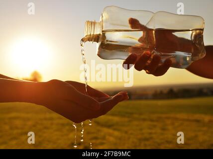 Die Hand, die das Trinkwasser aus der Flasche bei Sonnenuntergang in die Handfläche gießt. Stockfoto