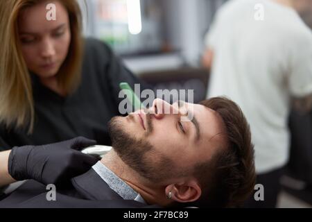 Young adult beautiful caucasian woman hairdresser cuts beard handsome man at modern barbershop Happy gay sitting chair men beauty salon. beard care. metrosexual. Female barber in black gloves serving Stockfoto