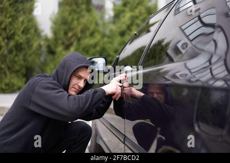 Auto jacking Dieb stehlen Auto brechen Tür kriminellen Job Einbrecher Hijacks Auto Dieb schwarz Balaclava Hoodie Versuch brechen in Fahrzeug Schraubendreher Straße Kriminalität Gewalt Gangster Räuber Auto Parken Stockfoto