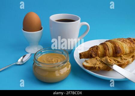 Frühstück: Croissant mit Erdnusspaste, schwarzer Kaffee und gekochtes Ei auf blauem Hintergrund Stockfoto