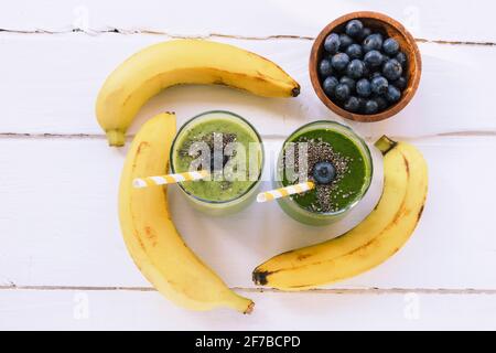 Zwei grüne Smoothies in Gläsern mit Trinkhalmen, Bananen und Heidelbeeren in einer Schüssel. Gesundes Lebensmittelkonzept. Weißer Holztisch Hintergrund. Draufsicht, flach liegend Stockfoto