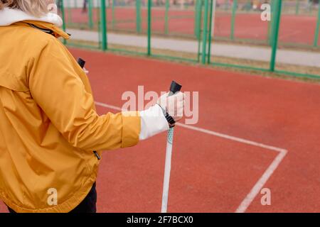 Eine ältere Frau in einer gelben Jacke hält im Stadion einen skandinavischen Stock in den Händen. Gesunder Lebensstil Stockfoto