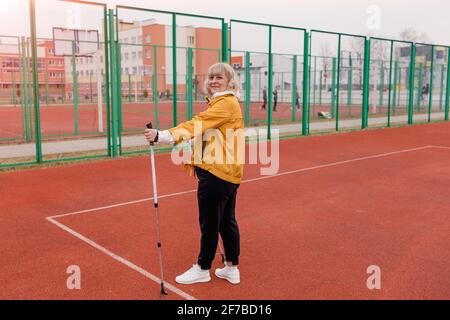 Eine ältere Frau in einer gelben Jacke hält im Stadion einen skandinavischen Stock in den Händen. Gesunder Lebensstil Stockfoto