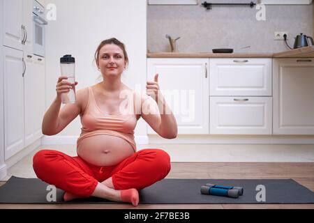 Fitness während der Schwangerschaft und die Vorteile von sauberem Wasser. Die Schwangere beim Sporttraining zu Hause zeigt den Daumen mit einer Flasche Wasser in sich Stockfoto