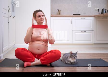 Sport während der Schwangerschaft mit einem Gummiband. Schwanger Frau macht Fitness-Training in der Küche zu Hause Stockfoto