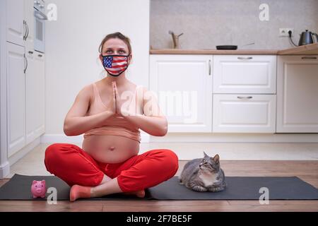 Fitnesskurse während der Schwangerschaft in der Gesichtsmaske zu Hause. Eine Schwangerin trägt bei einem Sporttraining in der Küche eine medizinische Maske mit dem Stockfoto