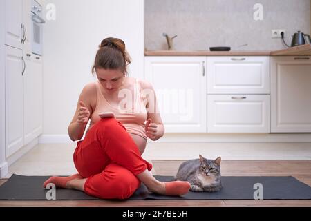 Sportliche Aktivitäten während der Schwangerschaft und Probleme Unterricht online. Eine Frau mit einem Telefon studiert Fitness über das Internet in einer Wohnküche Stockfoto