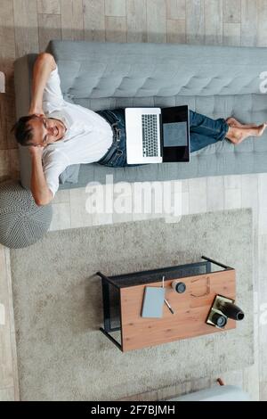 Glücklicher Mann mit einem Laptop, der auf der Couch liegt. Stockfoto