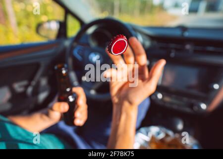 Junger Mann trinkt Bier im Auto und wirft Flaschendeckel aus. Konzentrieren Sie sich auf die rote Kappe in der Luft. Fahrer unter Alkoholeinfluss. Betrunkener Fahrer Stockfoto