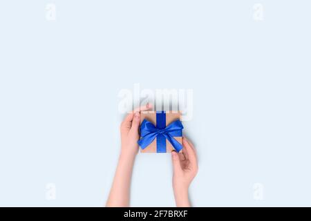 Weibliche Hände halten verpackte Geschenkbox auf einem blauen pastellfarbenen Hintergrund. Geschenk gebunden mit einem Band. Minimale Komposition mit Copyspace. Stockfoto