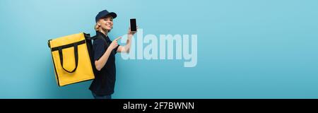 Fröhlicher Kurier mit Thermobeutel, der auf das Handy zeigt, mit leerem Bildschirm auf blauem Banner Stockfoto