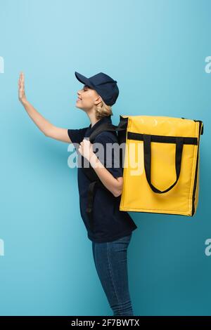 Seitenansicht des fröhlichen Kuriers mit der Hand winkender Thermotasche Auf blau Stockfoto