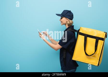 Seitenansicht eines lächelnden Kuriers mit einem Thermosack, der auf den Blick zeigt Handy auf blau Stockfoto