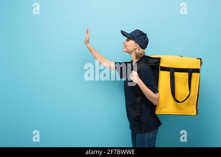 Seitenansicht des glücklichen Kuriers mit dem Thermorucksack, der die Hand winkt Auf blau Stockfoto