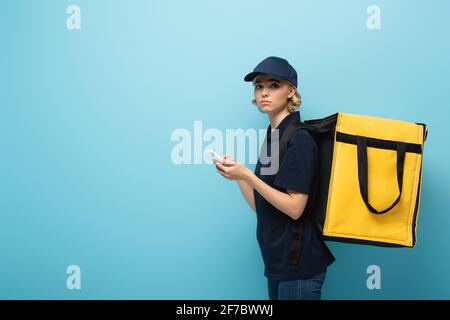 Verärgert Kurier hält Smartphone, während Thermotasche auf blau zu tragen Stockfoto