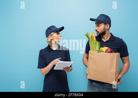 Der junge Kurier zeigte auf das digitale Tablet und überraschte fast den arabischen Kollegen Mit Lebensmitteln isoliert auf blau Stockfoto