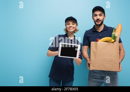 Fröhliche interracial Kuriere halten digitale Tablette mit leerem Bildschirm und Papiertüte mit Lebensmitteln isoliert auf blau Stockfoto