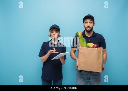 Verärgert Kurier zeigt auf digitale Tablette in der Nähe überrascht arabischen Kurier Papierbeutel mit Lebensmitteln auf blau isoliert halten Stockfoto