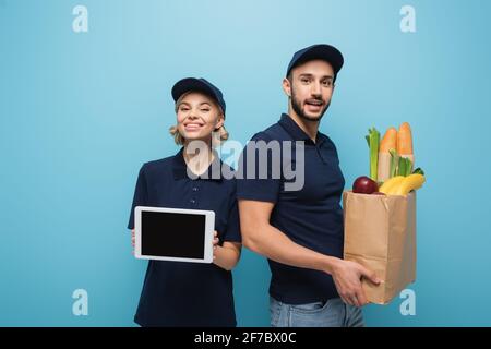 Fröhliche interracial Kuriere mit Essen in Papiertüte und digital Tablett mit leerem Bildschirm, der auf blau isoliert ist Stockfoto