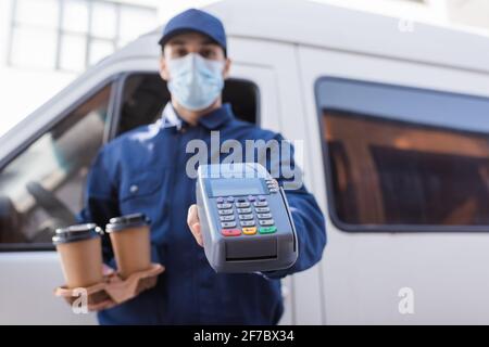 Selektiver Fokus auf Kaffee to go und Zahlungsterminal in Hände eines arabischen Kuriers in medizinischer Maske auf verschwommenem Hintergrund Stockfoto