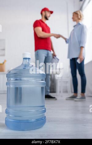 Selektiver Fokus der großen Flasche Trinkwasser in der Nähe von muslim Kurier und Frau schütteln sich die Hände auf unscharfem Hintergrund Stockfoto