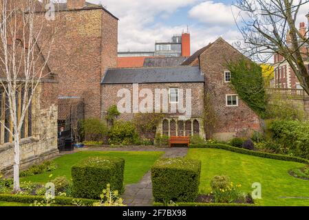 Ein kleiner Garten neben einem mittelalterlichen Gebäude. Eine Bank ist an einer alten Wand und vor einer Wiese aufgestellt. Ein weißer Baum und Hecken stehen im Vordergrund. Stockfoto