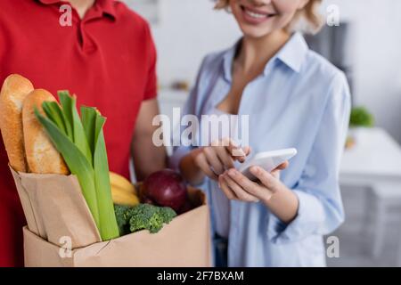 Teilansicht des Lieferers mit frischen Lebensmitteln auf Papier Tasche in der Nähe einer glücklichen Frau, die auf das Smartphone auf unscharfem Hintergrund zeigt Stockfoto