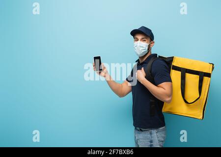 muslimischer Kurier in medizinischer Maske, zeigt Handy mit leerem Handy Bildschirm auf blau Stockfoto