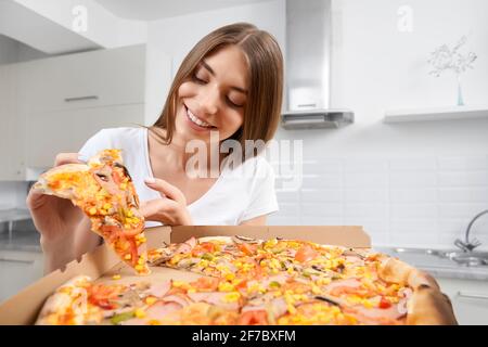 Nahaufnahme einer lächelnden Frau, die zu Hause eine große Pizza hält und ein Stück isst. Konzept des Essens leckere Speisen zu Hause. Stockfoto