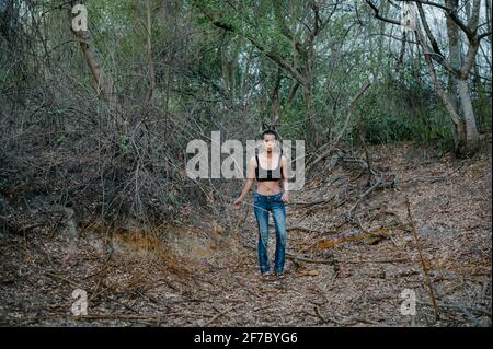 Lange Aufnahme einer jungen Asiatin mit kurzen Haaren, die allein in einem Wald steht, einen Sport-BH und blaue Jeans trägt Stockfoto