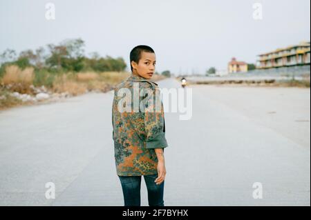 Junge Asiatin mit kurzen Haaren, die einen Sport-BH und eine Armeejacke trägt, geht weg, während sie über ihre Schulter auf die Kamera schaut. Stockfoto