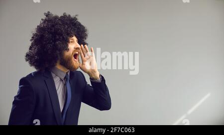 Lustiger Mann in lockiger Perücke, der laut auf grauer Werbung schreit Hintergrund des Kopierbereichs Stockfoto