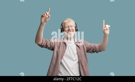 Fröhlicher Albino-Mann, der Musik in kabellosen Kopfhörern genießt, tanzt und die Finger über türkisfarbenem Studiohintergrund hebt Stockfoto