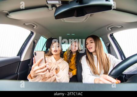 Drei glückliche Freundinnen gehen auf eine Reise. Frauen fahren im Auto, schauen auf das Telefon und lachen. Stockfoto